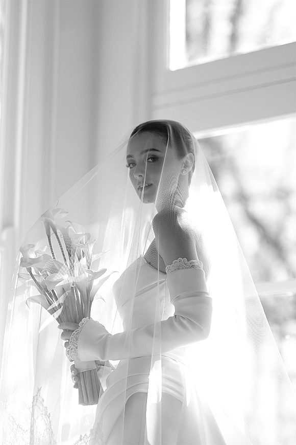 Bridal portrait in black and white of a bride under veil holding a calla lily bouquet by a large window, trees softly blurred outside