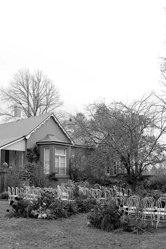 Ceremony setup with outdoor wedding ceremony seating, white chairs in rows framing aisle florals and greenery on a garden lawn by a house