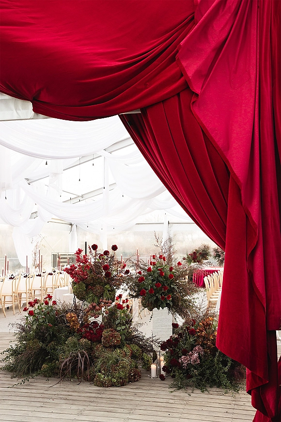 Reception decor with red wedding draping, burgundy florals, greenery, and taper candles on round tables beneath a white draped tent ceiling