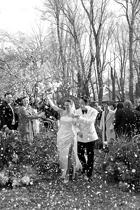 Wedding recessional as bride in strapless dress raises bouquet and walks with groom through confetti petals on a garden aisle, guests cheering