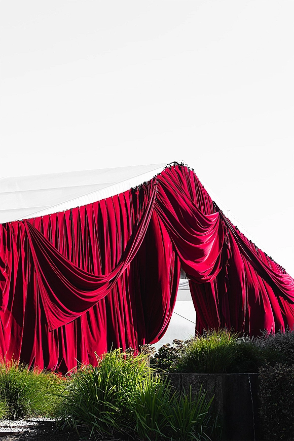 Ceremony backdrop with red draped wedding backdrop fabric swag under a tent canopy, set against ornamental grasses and a white sky
