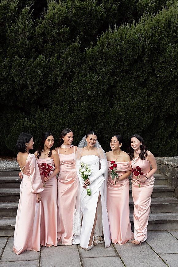 Bride with bridesmaids in blush satin dresses on stone steps, bride in strapless gown and veil holding calla lilies, others with red bouquets
