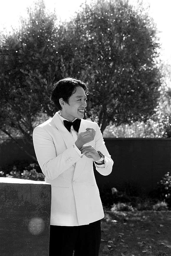 Groom portrait of a white tuxedo groom adjusting cufflinks, black bow tie and wristwatch visible, standing on a garden lawn near trees