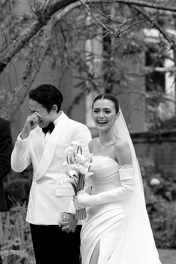 Ceremony moment as bride and groom holding hands, bride in strapless dress with cathedral veil and calla lily bouquet outdoors