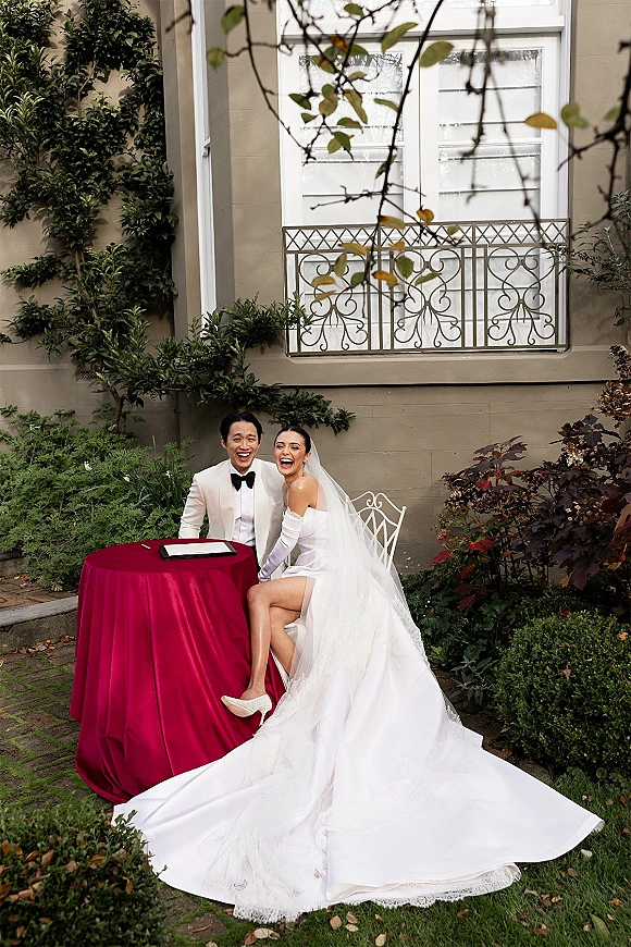 Couple portrait of a laughing bride and groom seated at a red sweetheart table, her long veil draped over white chairs in a garden courtyard