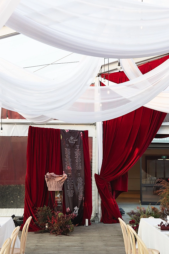 Wedding seating chart with a large seating chart sign framed by burgundy velvet drapes, string lights, and florals in a tent entrance