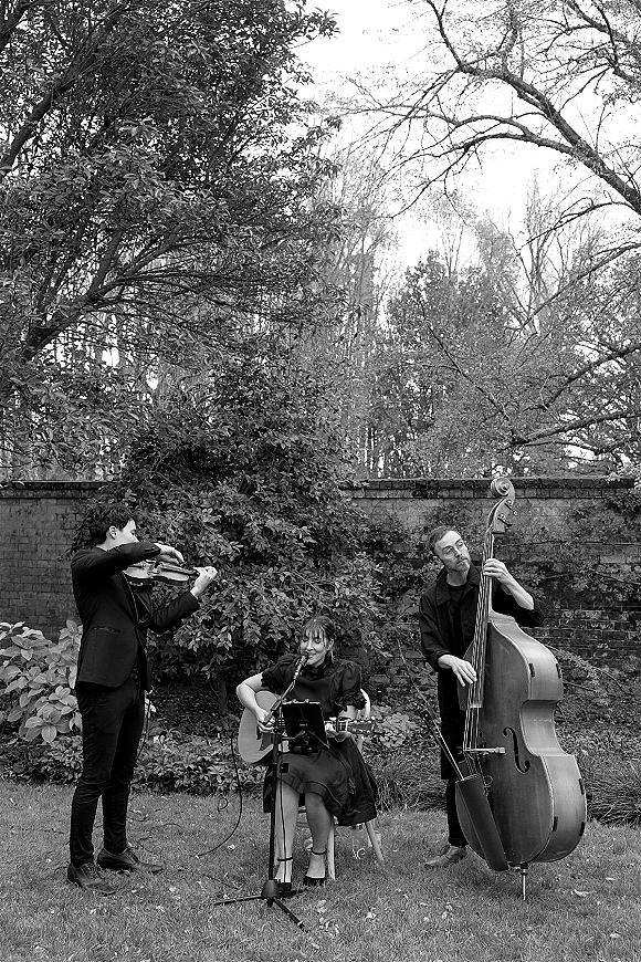 Wedding musicians with violin, acoustic guitar, and upright bass set up on a garden lawn by a brick wall and trees, with microphone stand