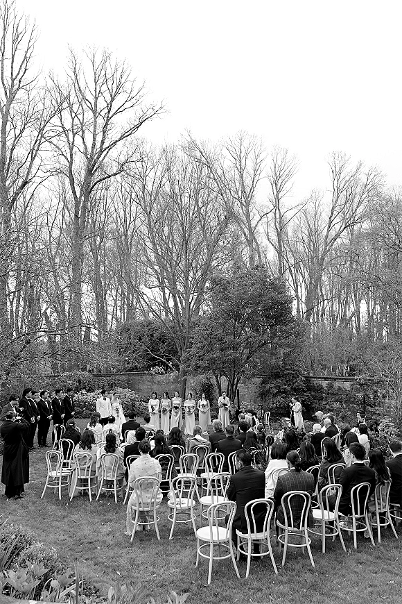 Outdoor wedding ceremony with bride, groom and bridal party lined up by white chairs on a garden lawn, framed by bare trees and stone wall