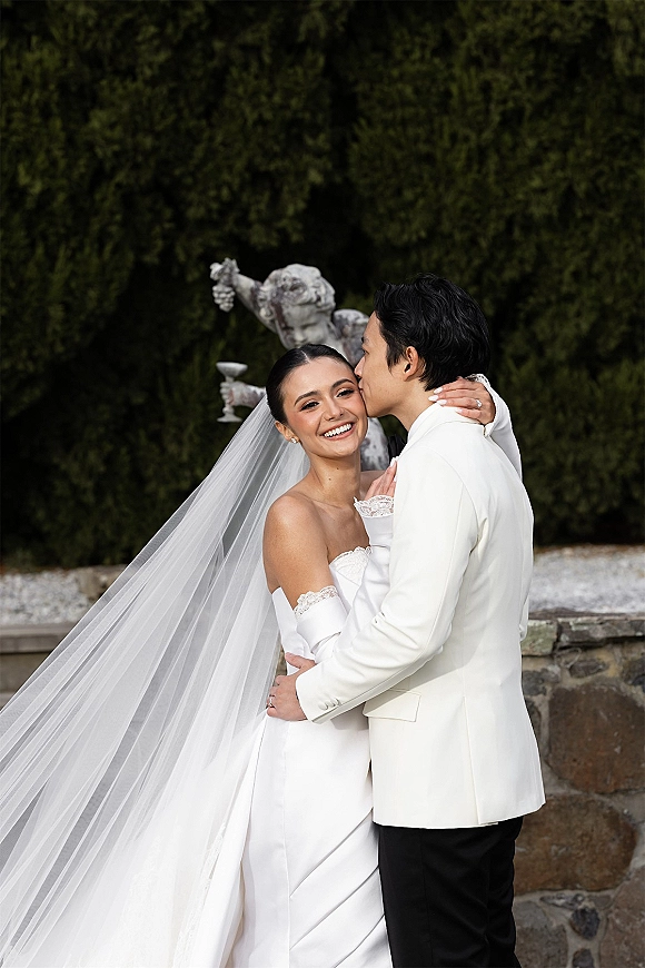Couple portrait of bride and groom hug as he kisses her cheek, cathedral veil flowing beside garden hedges, stone wall and cherub statue