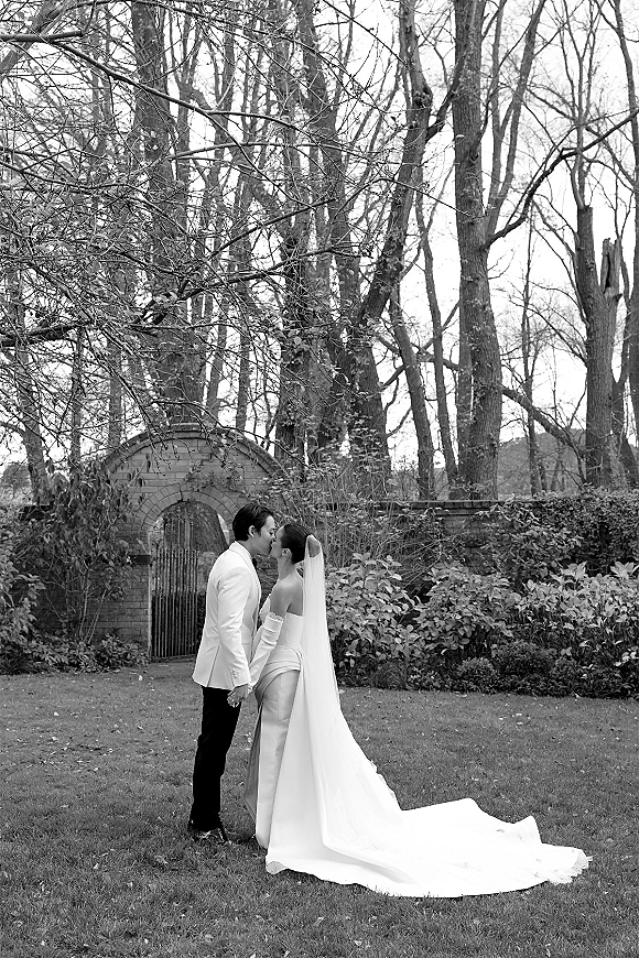 Wedding kiss portrait in black and white, bride and groom holding hands as her long train and veil trail across a garden lawn by stone wall and arched gate