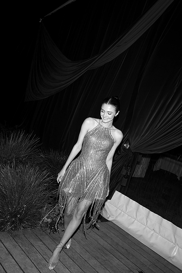 Wedding guest outfit in a sequin wedding guest dress with fringe hem, drop earrings and ankle strap heels on a wooden deck by draped fabric