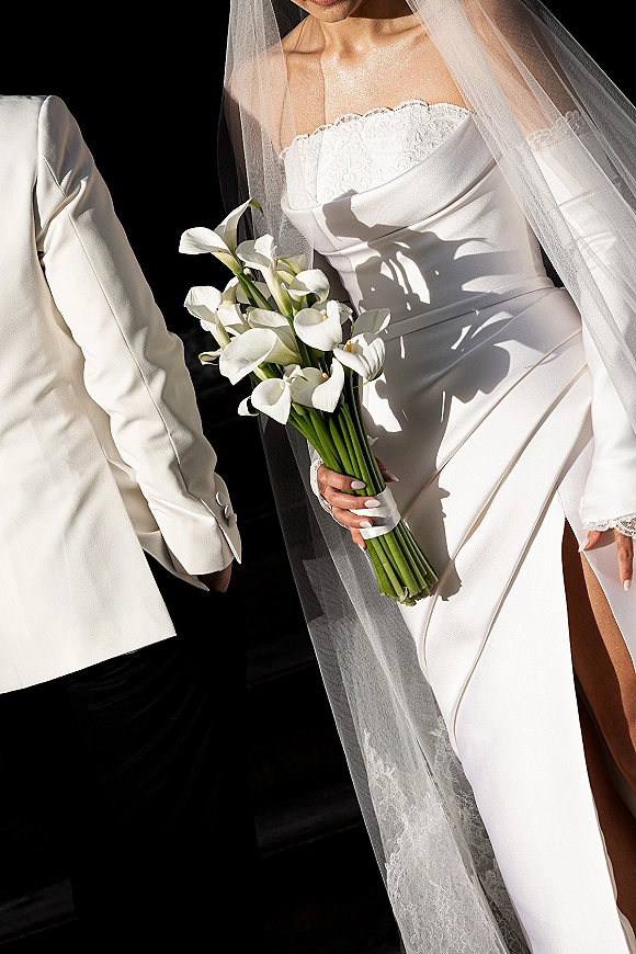 Bridal portrait with a calla lily bridal bouquet, showing strapless lace gown, veil, ring and manicure against a dark backdrop with shadow