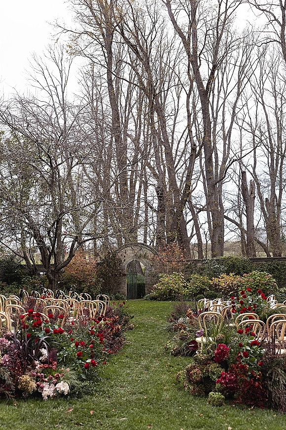 Ceremony aisle decor with red floral arrangements lining a garden lawn aisle, flanked by wood chairs and a stone archway backdrop