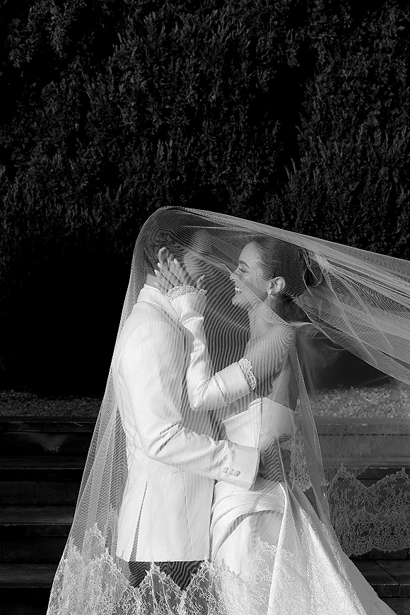 Wedding kiss portrait of bride and groom under veil, lace trim framing their faces on stone steps with hedges behind