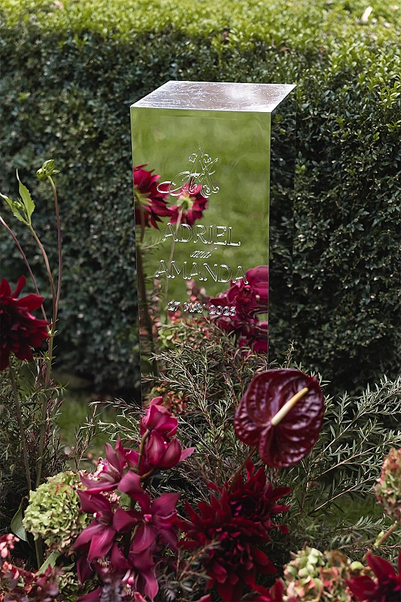 Wedding signage on a mirror wedding sign with engraved monogram and burgundy anthurium florals, set by a garden hedge on the lawn