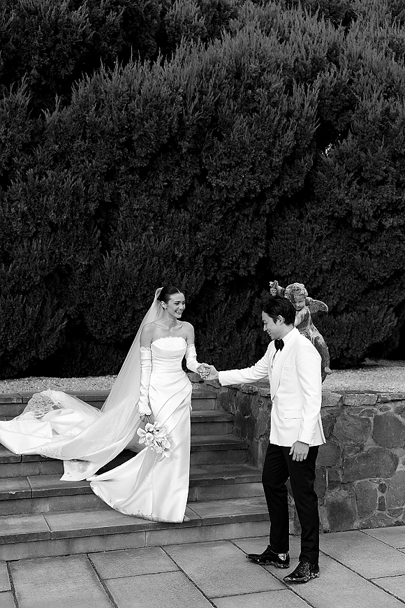 Couple portrait of bride and groom holding hands on stone steps, her strapless gown and veil trailing as he wears a white tuxedo