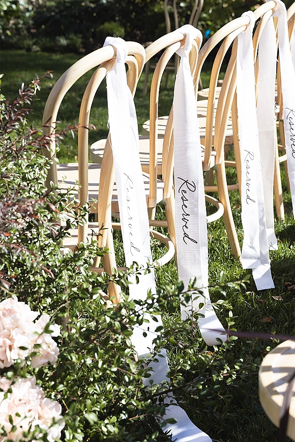 Ceremony seating with reserved ceremony chairs, wood chairs and white ribbon signs on a sunlit lawn with garden shrubs behind