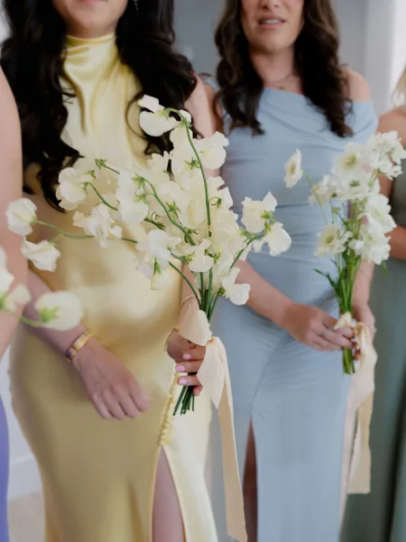 Bridesmaid bouquets of white sweet pea bouquet blooms held by bridesmaids in pastel dresses, finished with long ribbon streamers indoors
