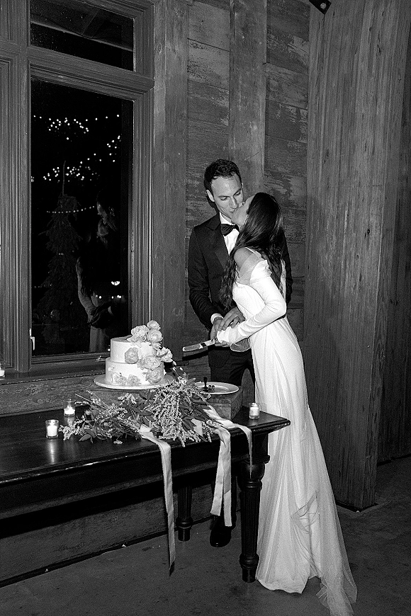 Wedding cake cutting with bride and groom kissing beside a two-tier cake, candles and champagne flutes on a wooden table by a window at night