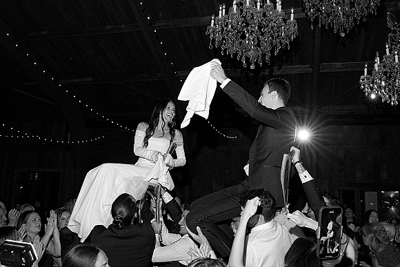Hora dance at a Jewish wedding hora as bride and groom are lifted on chairs, guests cheering under chandeliers and string lights in a hall