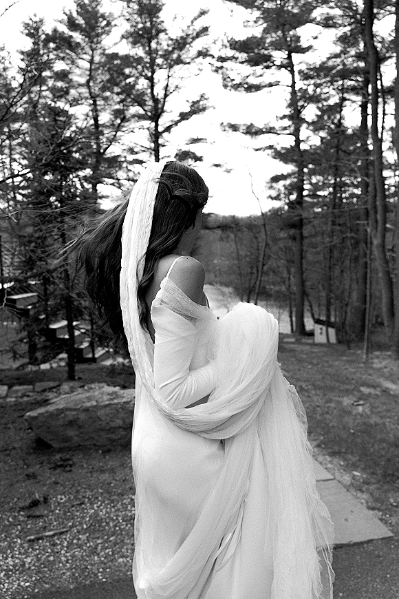 Bridal portrait of a bride from behind in a wedding dress, holding her train as her veil blows along a forest hillside path