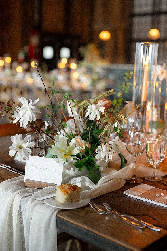 Reception tablescape with wedding table centerpiece of white blooms and greenery, tall cylinder candles, and place cards in a dim warm-lit room