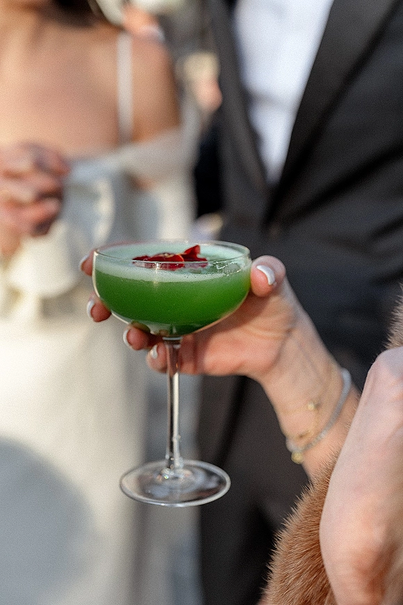 Wedding cocktail in a coupe glass, green and foamy with rose petal garnish, held by manicured hands as the couple blurs behind