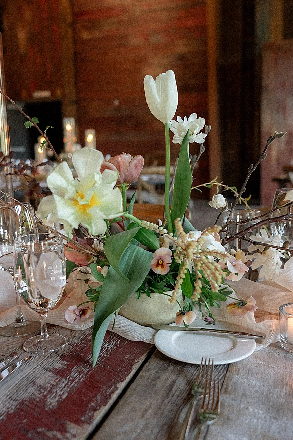 Reception tablescape with wedding table centerpiece of tulips and pansies in a ceramic bowl, with candlelit place settings in rustic wood interior