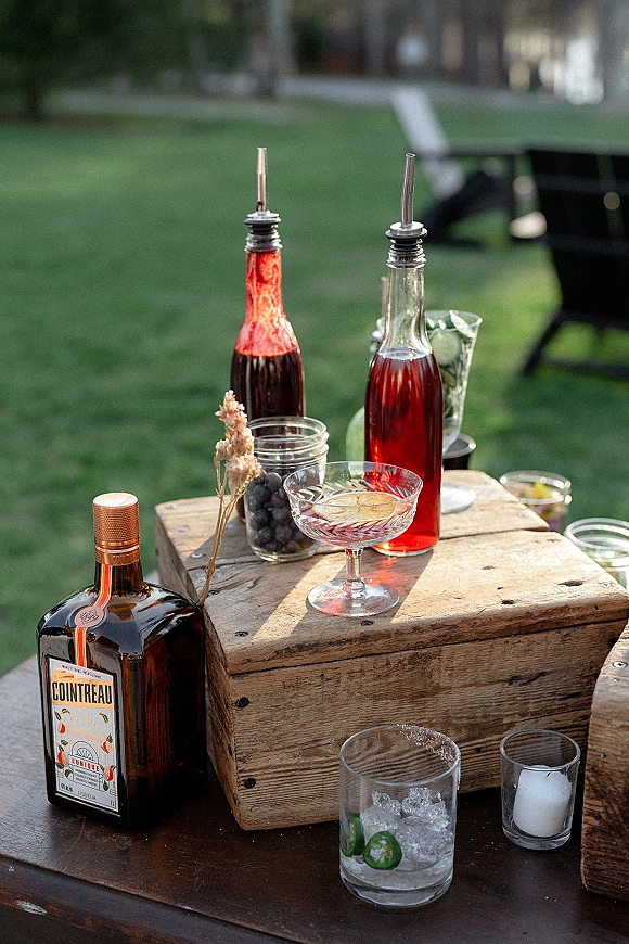 Wedding bar setup with liquor bottles, bitters, coupe and rocks glasses, limes, blueberries, cucumber slices and ice on a lawn picnic table
