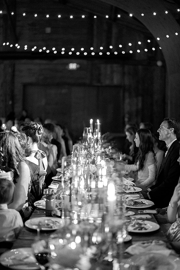 Reception dinner with a long banquet table wedding setup, taper candles and floral centerpiece glowing under string lights in a rustic barn interior