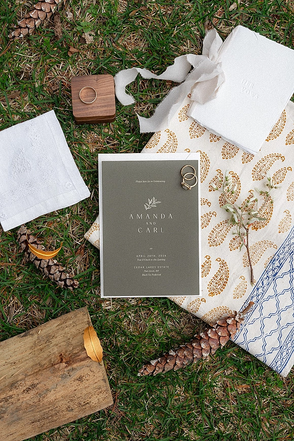 Invitation suite wedding invitation flat lay with envelope, rings in a wood box, ribbon, and pinecones on fabric over a grass lawn