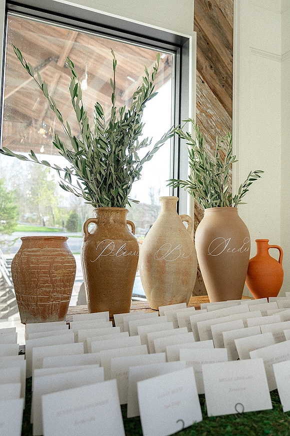 Escort card display with wedding escort cards in calligraphy, ceramic vases and olive branches on a wooden table by a large window.