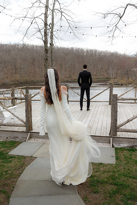 Wedding first look as bride approaching groom from behind, long-sleeve gown and veil trailing on a string-lit dock by the lake