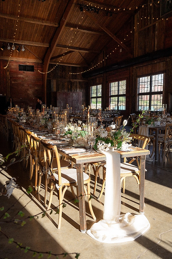 Reception tablescape with rustic barn reception details, wood farm tables, white florals and greenery, taper candles under string lights in a barn
