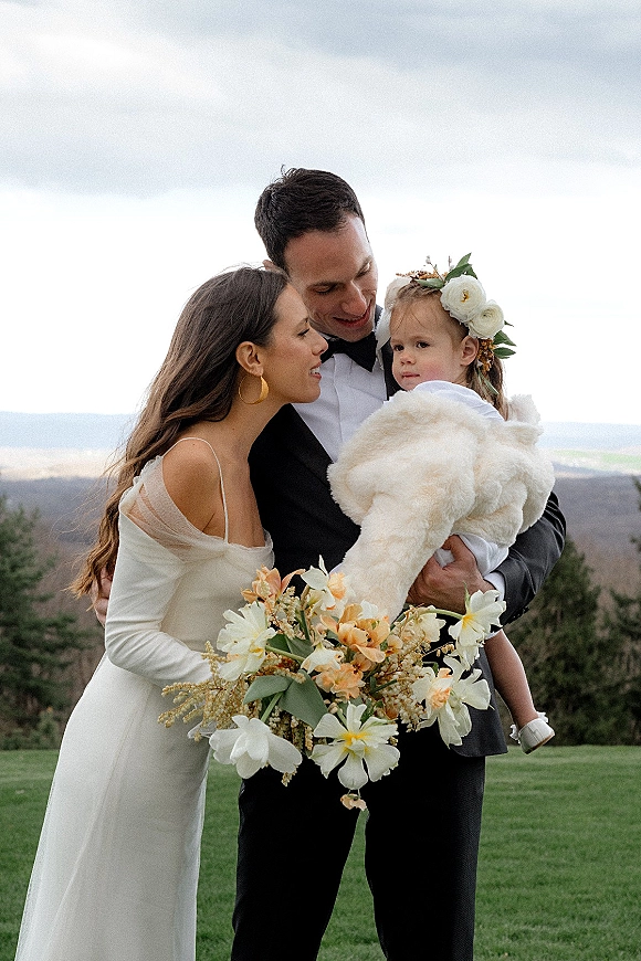 Family wedding portrait of bride and groom holding their child, bride with bouquet and floral crown, mountains under cloudy sky behind