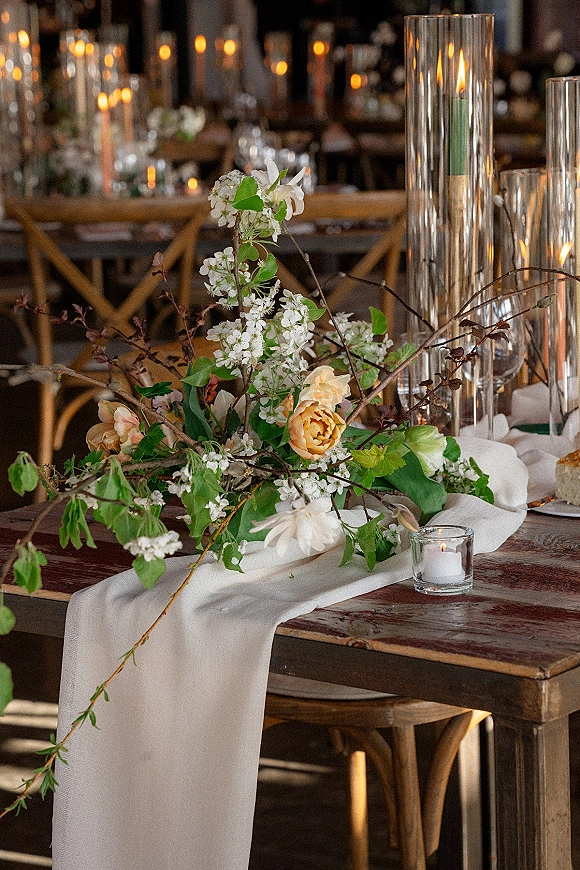 Wedding tablescape with rustic wedding table decor featuring peach roses, white blossoms, taper candles and glass vases on a wood table with cross back chairs