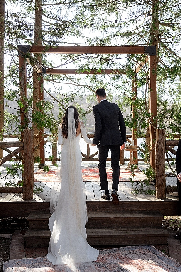 Wedding recessional as bride and groom walk away hand in hand under a greenery garland arch with string lights by a lakeside deck