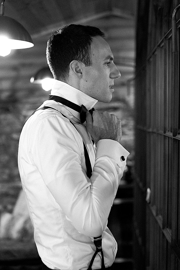 Groom portrait with black bow tie and suspenders, adjusting his tie in a wood-paneled room lit by a warm lamp