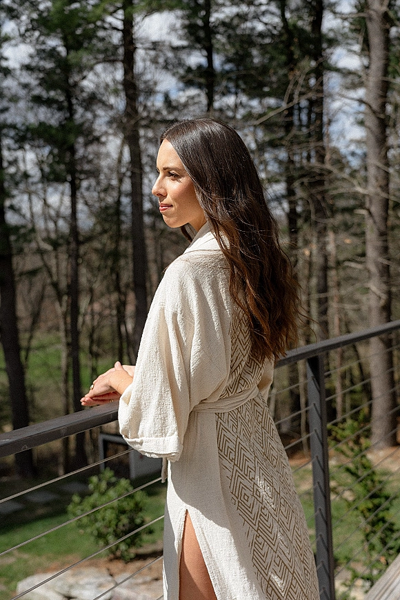 Bridal portrait of a bride in robe with a waist tie belt, standing on an outdoor balcony railing with forested hillside behind her