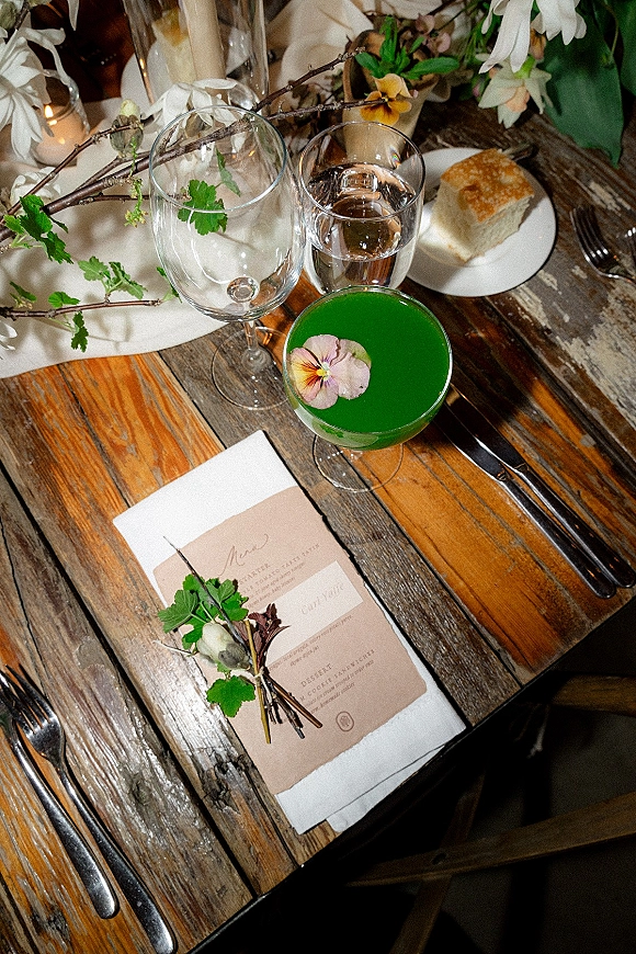 Reception tablescape on a rustic wedding tablescape wood table with menu card, candlelight, floral centerpiece, and green cocktail with edible flower garnish