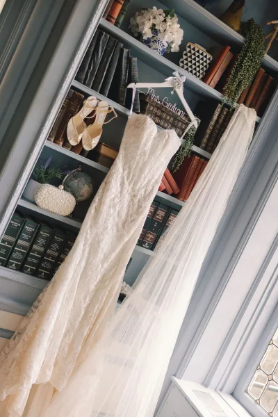 Wedding dress in lace hanging on a personalized hanger with tulle veil, shoes and pearl clutch on a built-in bookshelf by a window