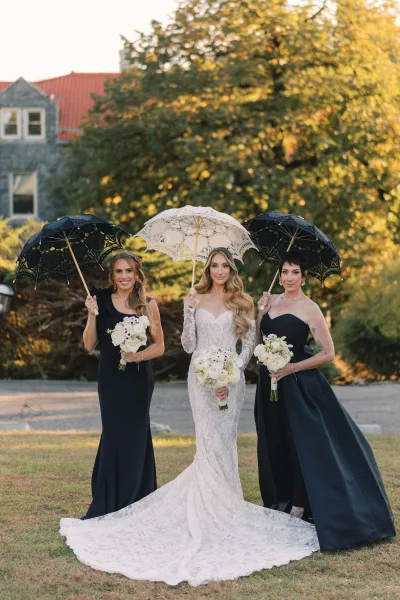 Bride with bridesmaids portrait showing lace-sleeved wedding dress and bouquets, with bridesmaids holding black parasols on a lawn by a stone building
