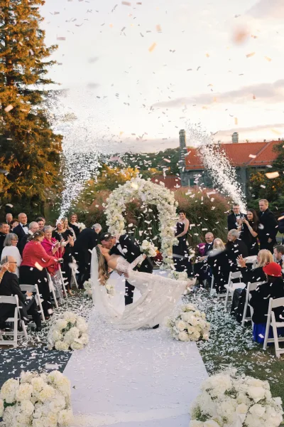 Ceremony kiss moment as bride and groom dip-kiss beneath a white floral arch, wedding recessional confetti raining at sunset garden aisle