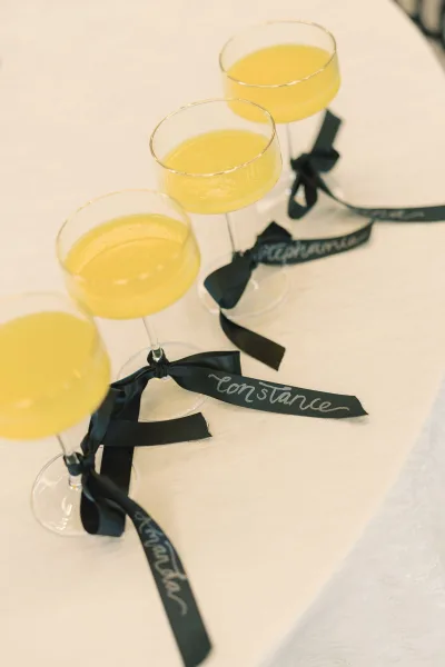 Wedding escort drinks arranged as an escort card drinks display with yellow cocktails in champagne coupes, black ribbon bows, and handwritten name tags on white linen tablecloth