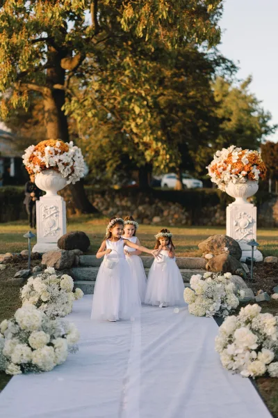 Flower girl processional with flower girls walking down aisle in white dresses, carrying baskets beside a white aisle runner outdoors