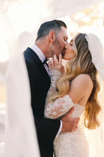 Wedding kiss portrait of bride and groom kissing under a lace veil, her ringed hand on his face as he wears a black tuxedo in soft light