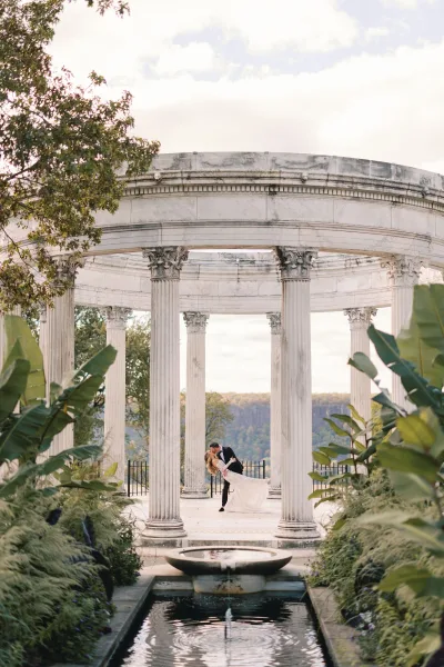 Wedding kiss portrait of bride and groom in a dramatic dip kiss, veil flowing beside a fountain at a stone rotunda with columns and trees