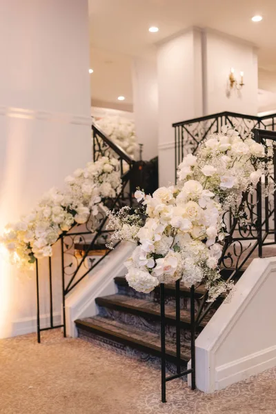 Wedding staircase florals drape a black wrought iron railing, with white roses, hydrangeas and orchids on an elegant indoor stairway