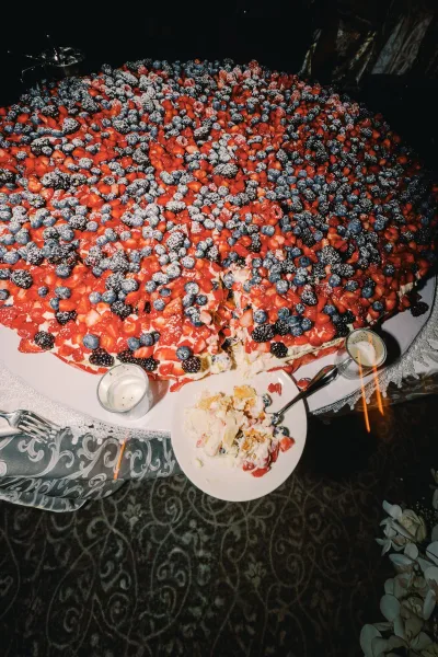 Wedding cake topped with mixed berries and powdered sugar on a lace tablecloth, surrounded by dessert plates and votive candles in dim light
