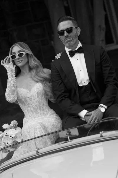 Couple portrait in a vintage convertible car, bride in lace dress and groom in tuxedo wearing sunglasses, building facade behind them